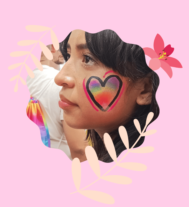 Un primer plano enmarcado de una mujer joven con cabello oscuro, vista de perfil, mirando hacia la izquierda. La parte superior de su mejilla derecha presenta una pintura facial detallada: un corazón colorido